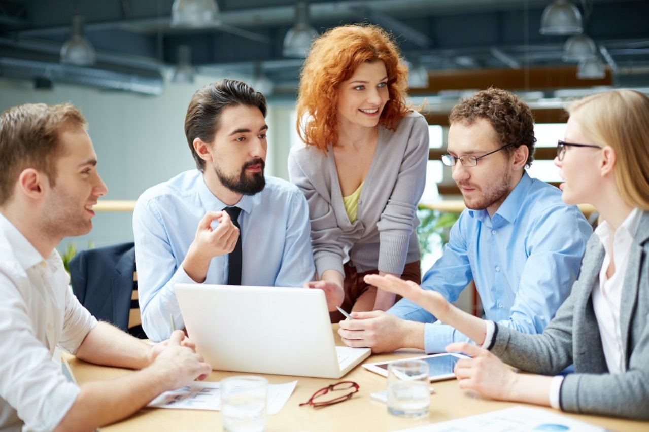 Group of professionals gathered around a table in a modern office, engaged in a collaborative discussion with a laptop and documents in front of them, suggesting a team meeting or brainstorming session.