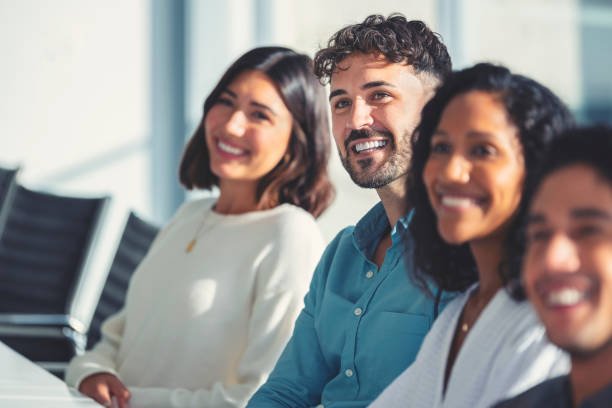 Group of people listening to a presentation. The are at a seminar or meeting sitting at a table. They are all looking to the front of the room. Men and women dressed in business attire. Multi-cultural group with African, Latino and Caucasian