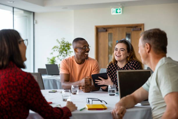 Medium shot of a mixed ethnic group of business professionals at a networking conference/meeting room in the North East of England. They are sitting at a table using digital tablets/laptops laughing together.