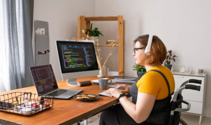 A woman using a wheelchair sits at a desk with two computer screens, wearing headphones while working on coding tasks in a modern home office
